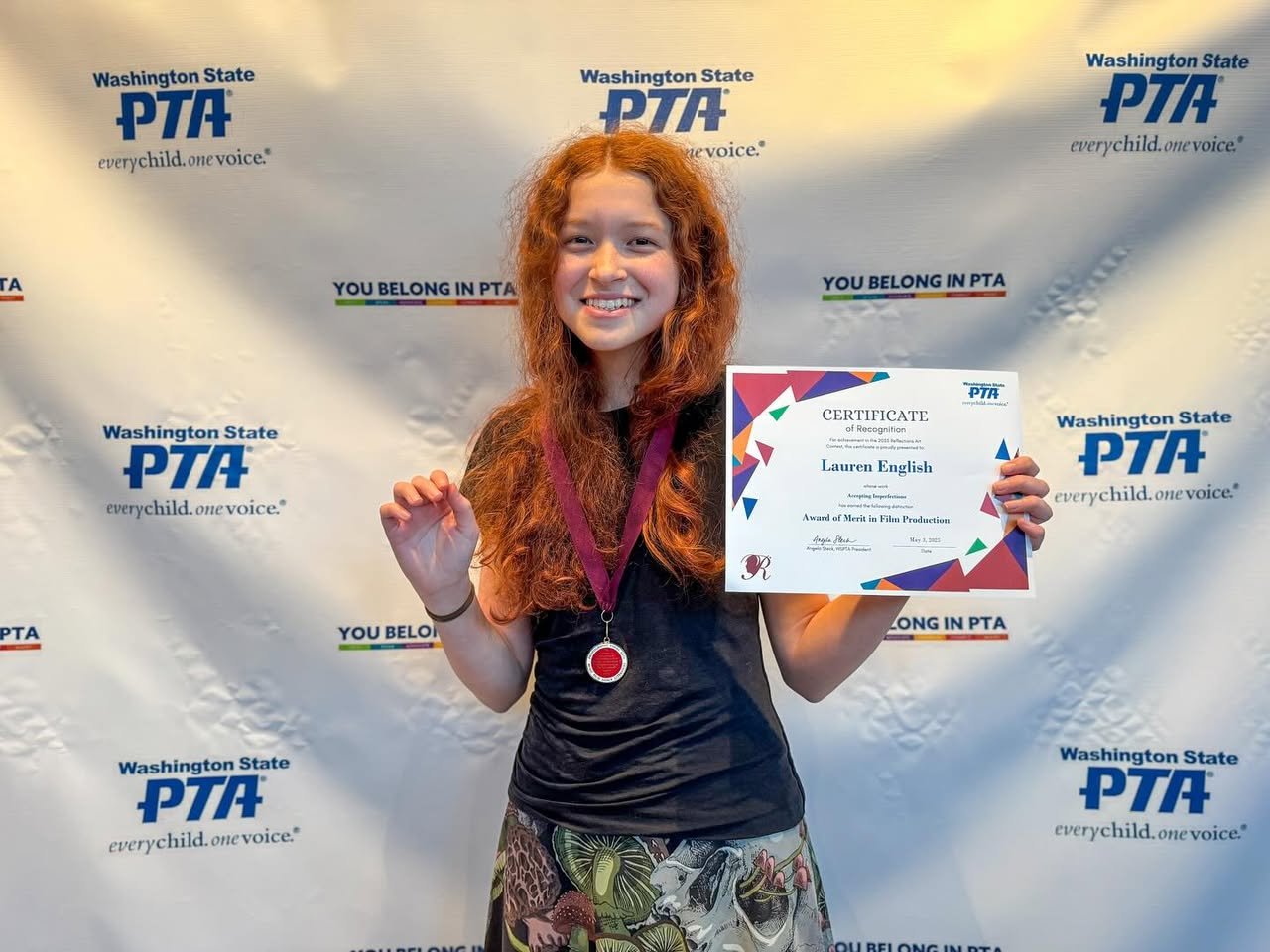 Washington State PTA Reflections A young person with long red hair smiling, wearing a medal, and holding a certificate in front of a Washington State PTA backdrop.