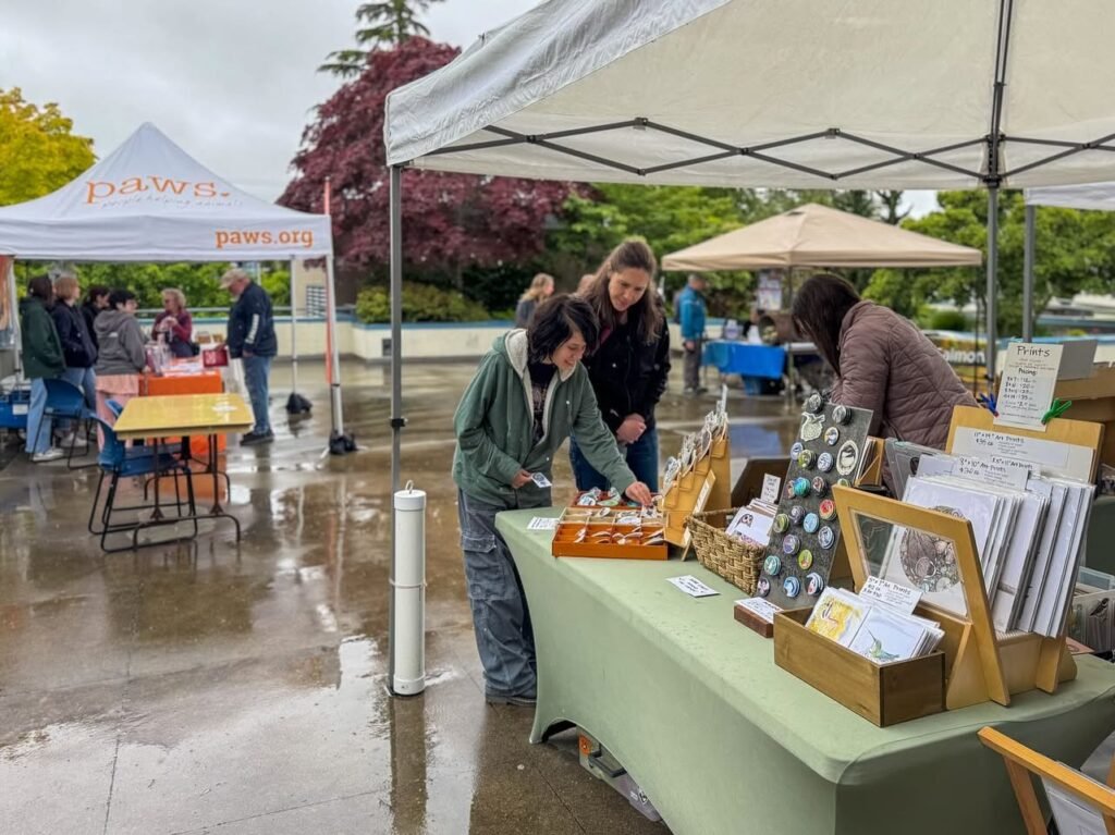 Browsing local market stalls under a tent in the rain. Wood-framed prints are displayed for sale at an outdoor event.