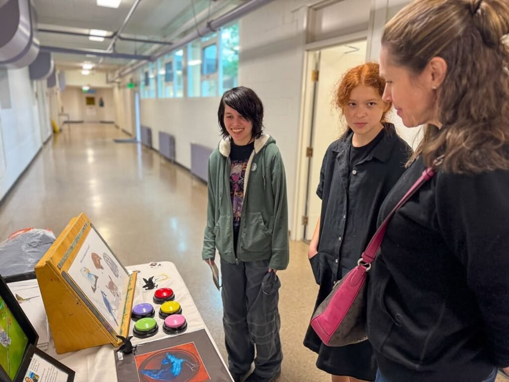 Students gathering around a science table with colorful buttons and an informational display in a school hallway, indicating an educational display.