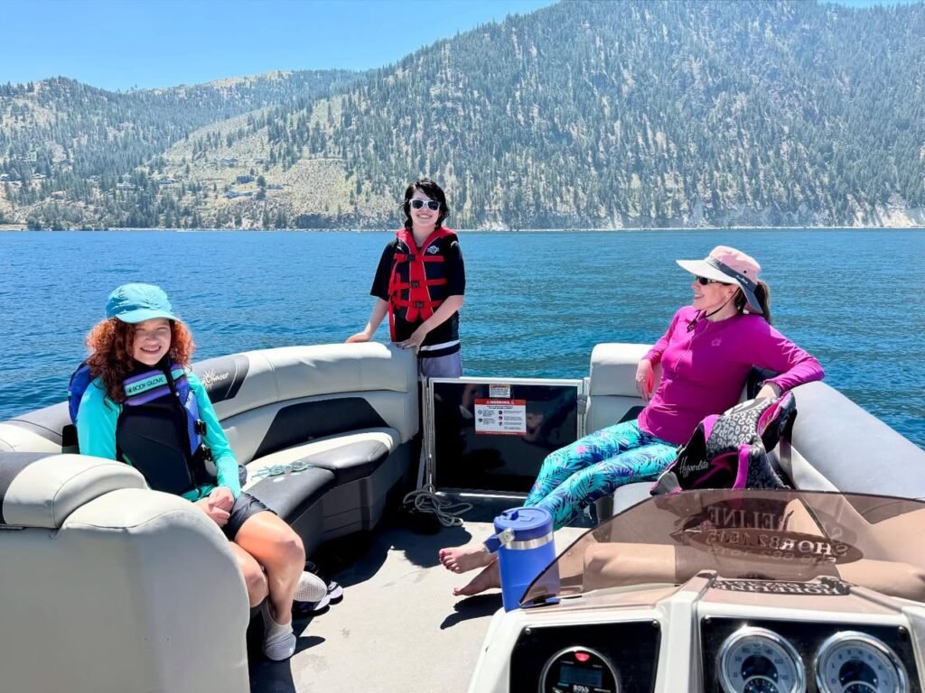 A group of people relax on a boat in a picturesque lakeside setting, with mountains in the background.
