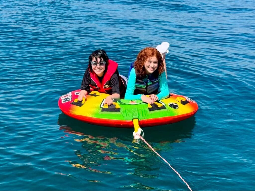 Two people float on a colorful inflatable raft on a calm blue lake, wearing life jackets and enjoying a sunny day on the water.