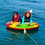 Two people float on a colorful inflatable raft on a calm blue lake, wearing life jackets and enjoying a sunny day on the water.