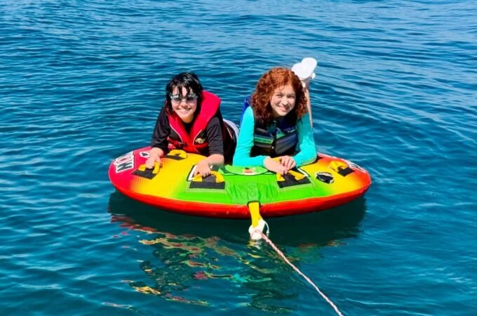 Two people float on a colorful inflatable raft on a calm blue lake, wearing life jackets and enjoying a sunny day on the water.