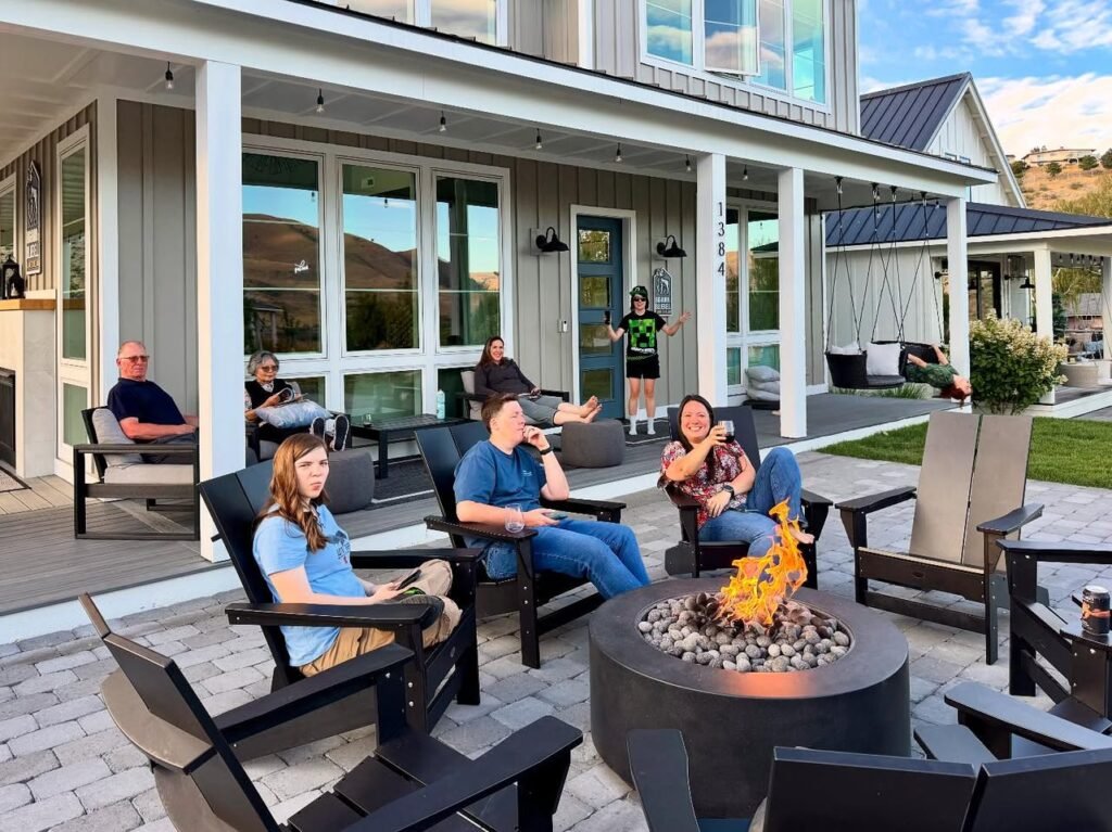 A group of people relaxing in outdoor chairs around a burning fire pit in front of a modern home with large glass windows.