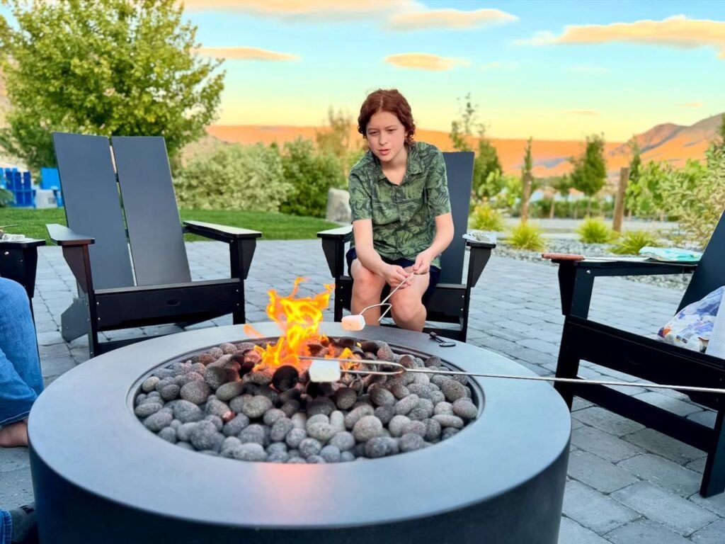A person sits by a gas fire pit roasting s'mores on a patio during sunset. Views of desert-like mountains and lush trees surround the paved outdoor space.