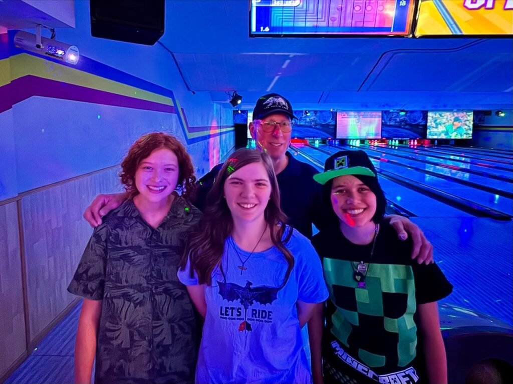 A family group poses happily in a bowling alley lane. The background features bowling lanes, score displays, and colorful lighting.