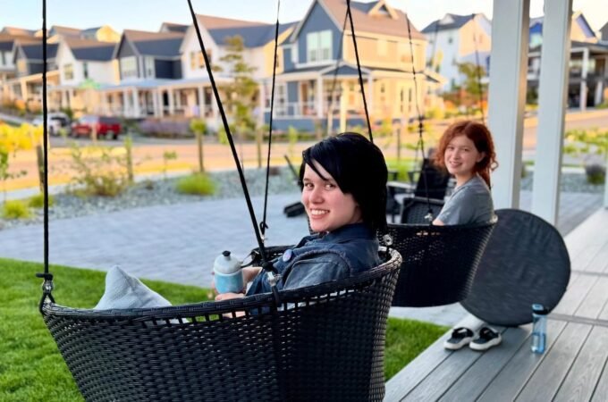 Two people relaxing in black hanging patio chairs on a wooden deck surrounded by well-maintained urban homes.