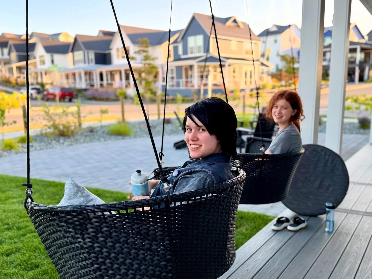 Summer in Chelan Two people relaxing in black hanging patio chairs on a wooden deck surrounded by well-maintained urban homes.