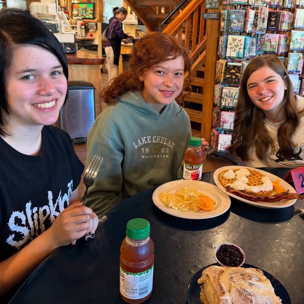 Three smiling people at a cafe table with breakfast plates and juice bottles, with a bookshelf in the background.