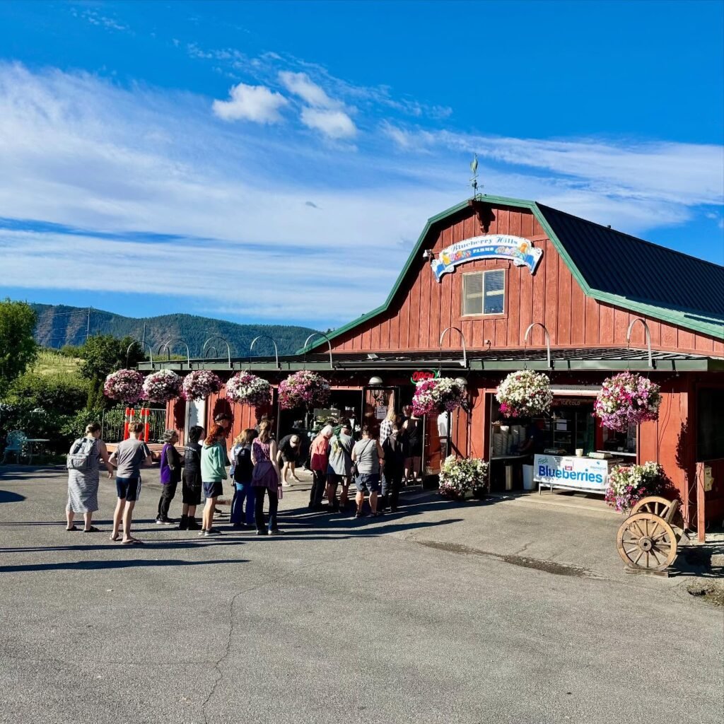 A red barn-style building with a green roof, featuring flower baskets and a group of visitors, set against a backdrop of rolling hills under a blue sky.