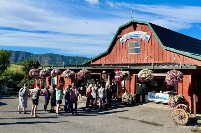 A red barn-style building with a green roof, featuring flower baskets and a group of visitors, set against a backdrop of rolling hills under a blue sky.