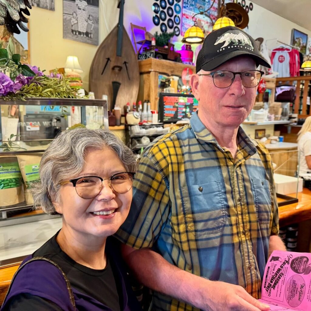 An older couple stands together in a vibrant, eclectic gift shop filled with colorful merchandise. The background features wooden artifacts, bright lighting, and floral displays.