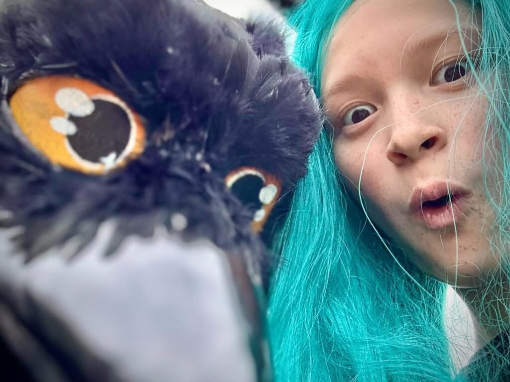 A person with bright turquoise hair poses playfully next to a detailed owl mascot.
