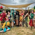A group of costumed garden gnomo performers poses together on a grassy field under a canopy. The costumes are colorful and whimsical, matching characters from Mary Poppins.