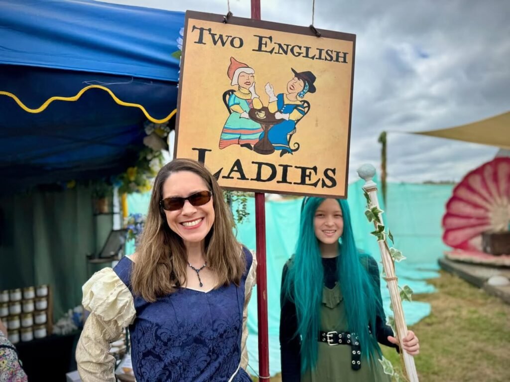 Two people pose beneath a sign for Two English Ladies at an outdoor folk festival. They stand in front of a blue tent with market goods visible nearby.