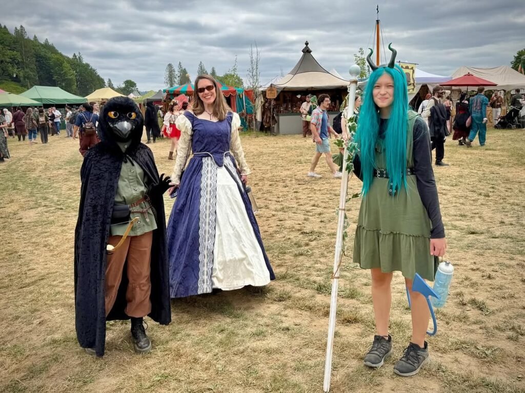 Enthusiasts in elaborate costumes and masks gather at an outdoor Renaissance fair. Colorful tents and a medieval-style structure are visible in the background.