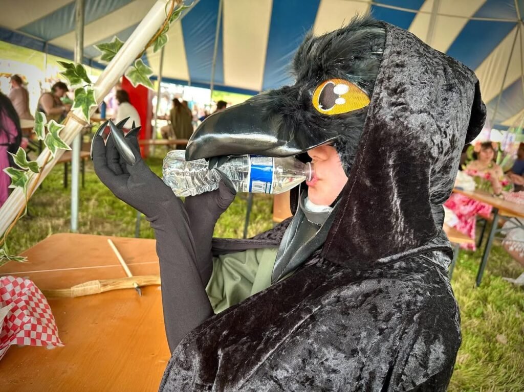 Person in a raptor costume eating under an event tent. The striped tent and surrounding decorations suggest a lively gathering.
