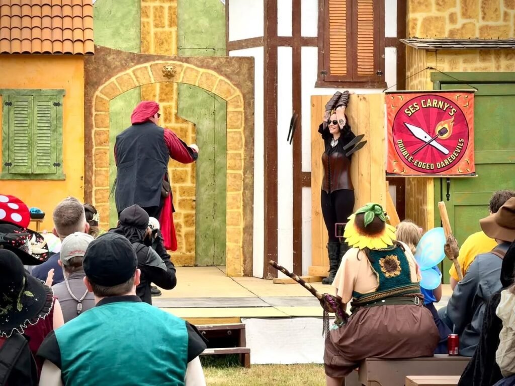 Actors in costume perform in front of a colorful medieval-style building. A sign reads "Sir Cerryl's Street Sports Services."