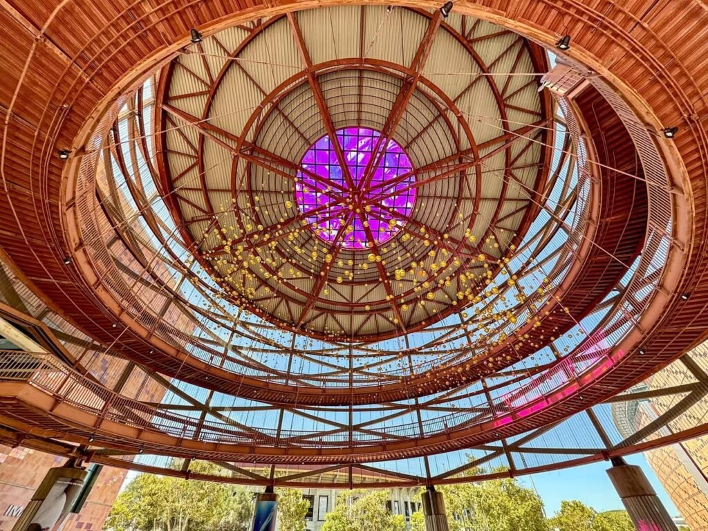 An architectural view of a wooden circular ceiling featuring a central violet glass panel. A lower section reveals curved wooden beams and translucent panels allowing natural light to pass through.