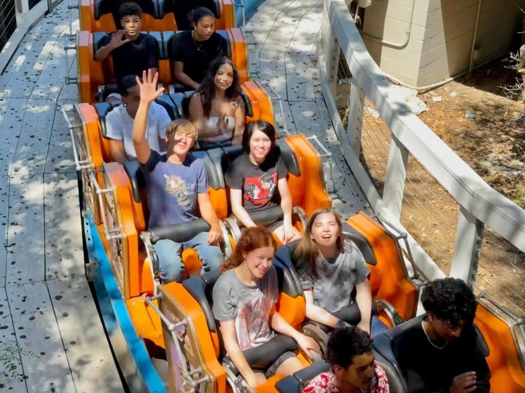 A group of people riding a roller coaster with orange seats. Some riders are waving, showing excitement.