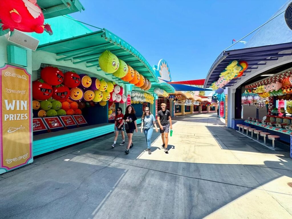 A sunny walkway at a colorful theme park feature with people strolling past a 'Win Prizes' booth.