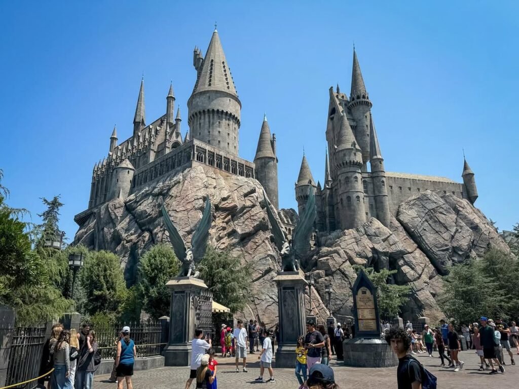 A majestic, multi-tiered castle with pointed turrets, built into a rocky cliffside. Visitors walk through the plaza, taking photos against a clear blue sky.