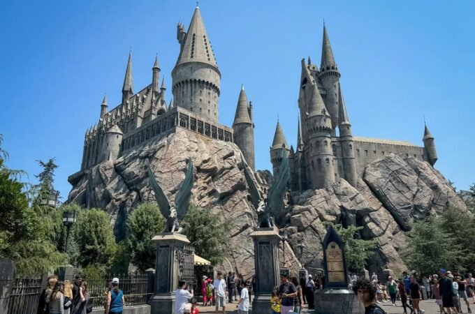 A majestic, multi-tiered castle with pointed turrets, built into a rocky cliffside. Visitors walk through the plaza, taking photos against a clear blue sky.