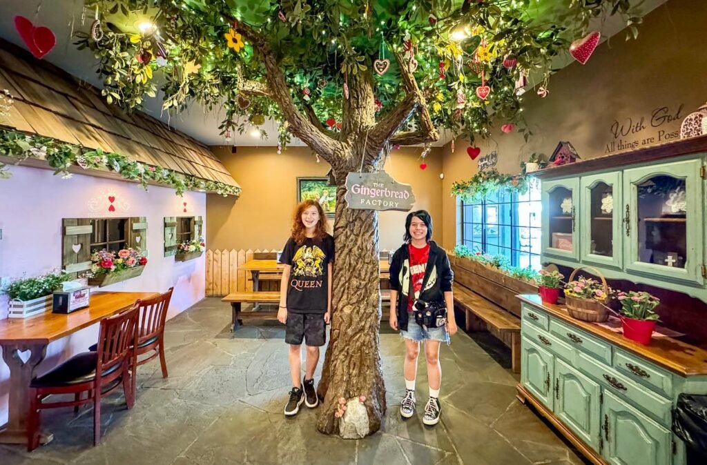 Two individuals stand next to a large decorative tree in a whimsical indoor setting with rustic furniture and cabinet details.