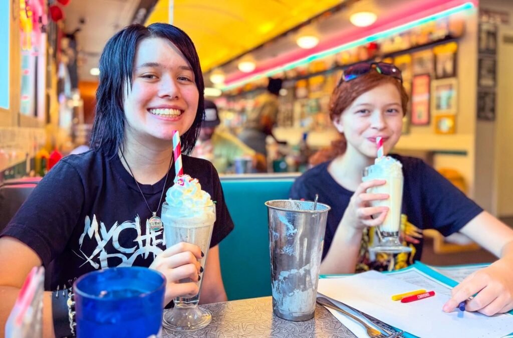 Two people enjoy cream sodas in a retro-style diner with colorful neon décor.