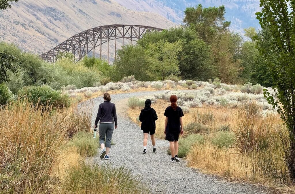 A group walks along a gravel path through dry grasslands, with a historic steel bridge and mountain backdrop visible.
