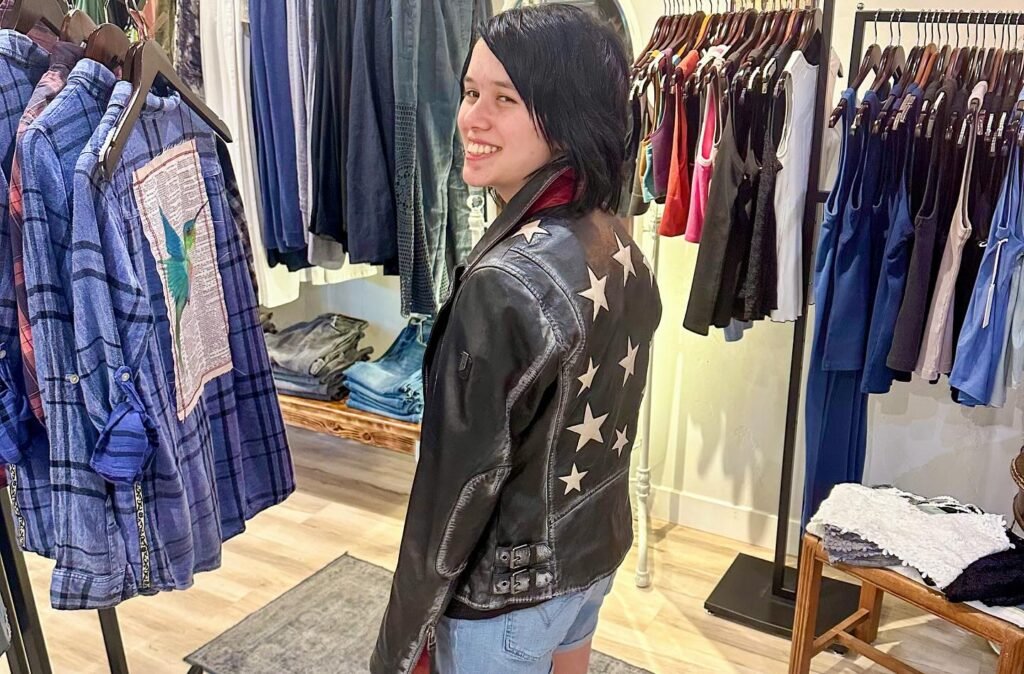 A person wearing a black leather jacket with white star details, surrounded by racks of clothing in a shop.