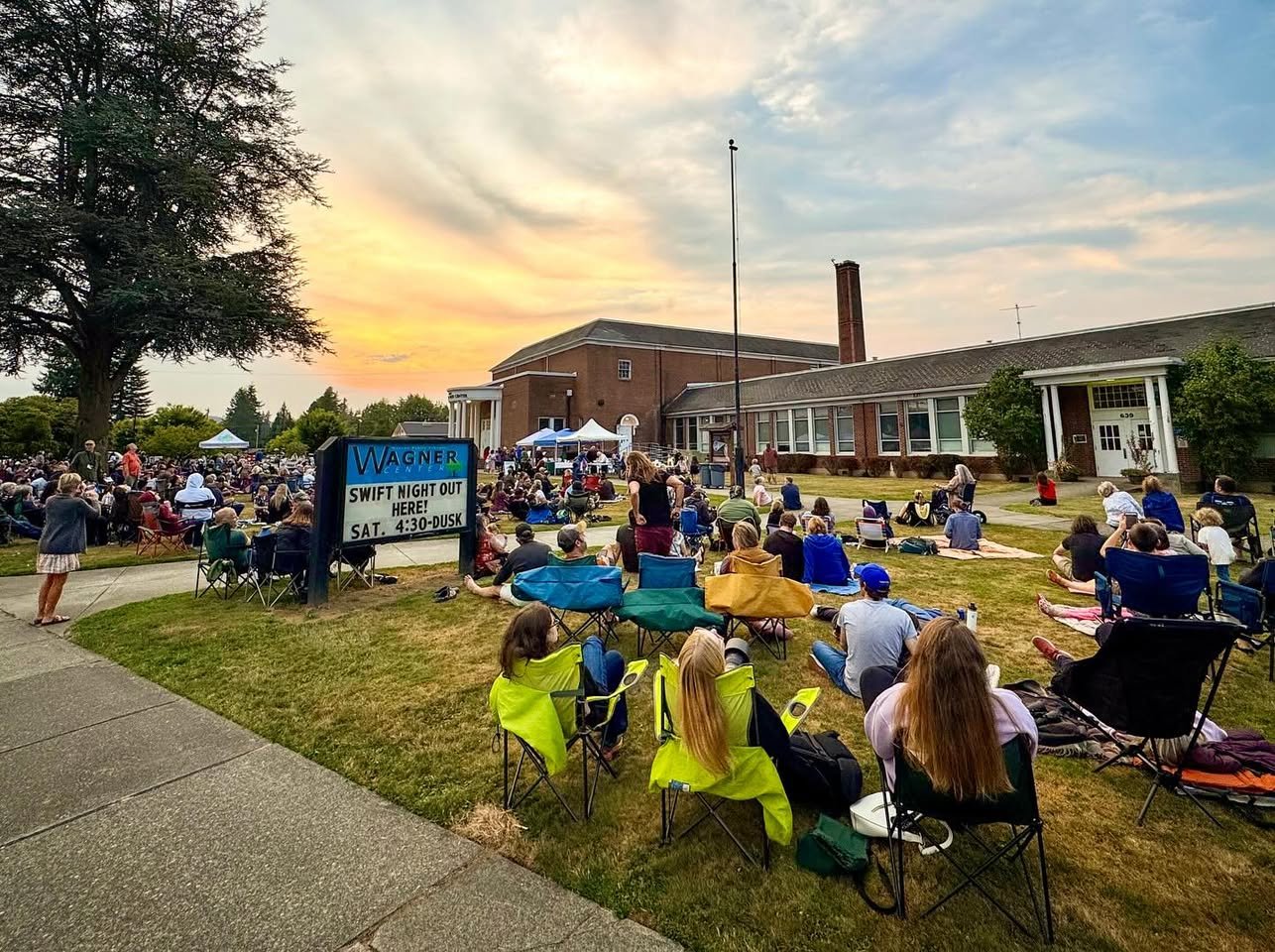 Swifts Night Out A crowd of people sits on lawn chairs in the grass toward a brick building during a golden hour sunset. People are gathered on a lawn with some standing near a sign.