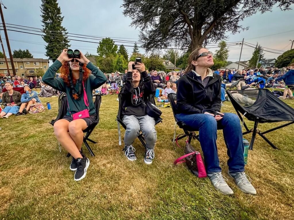Three people seated in folding chairs on a grassy field looking up at the sky during an outdoor event. A large crowd is gathered on the lawn in the background beneath tall evergreen trees.