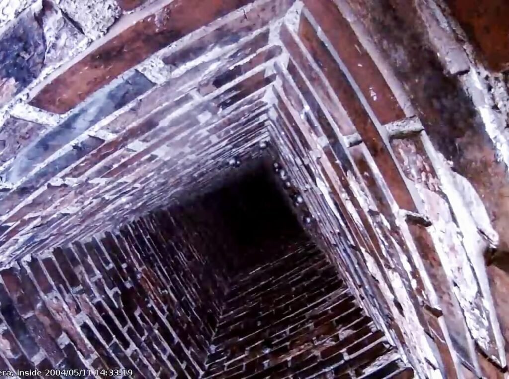 A view looking down the interior of an old brick chimney flue with a dark passage and textured masonry walls.