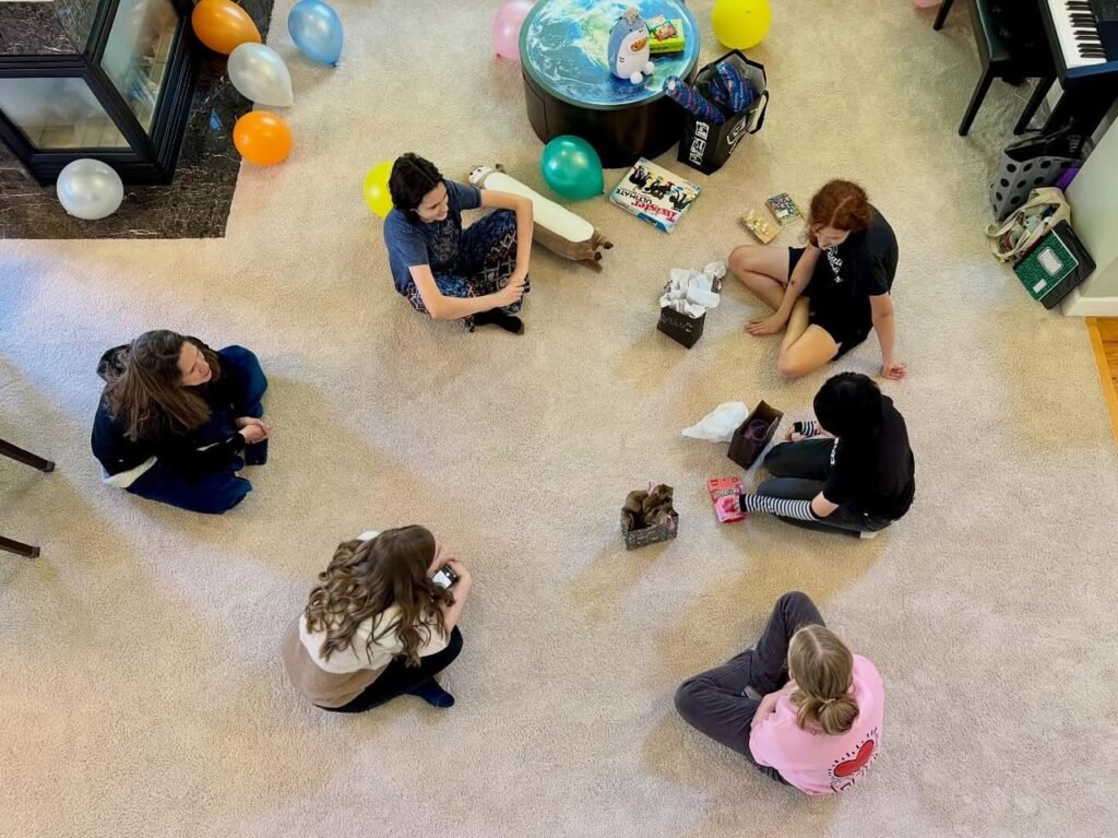 Children sitting in a circle on the floor with a globe and colorful balloons in the background.