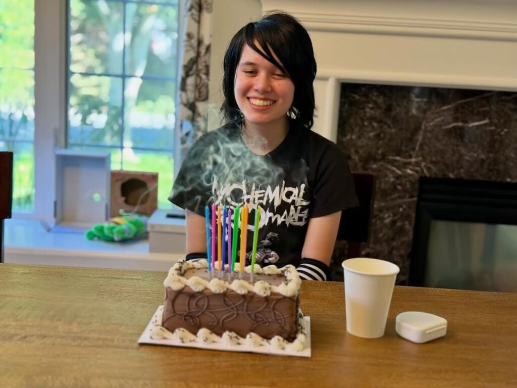 A person seated at a wooden table with a decorated birthday cake and colorful candles. The cozy scene includes a window with a view of greenery outdoors.