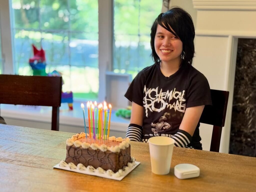 A person smiling beside a birthday cake with lit candles on a wooden table. The background shows a bright window and indoor setting.