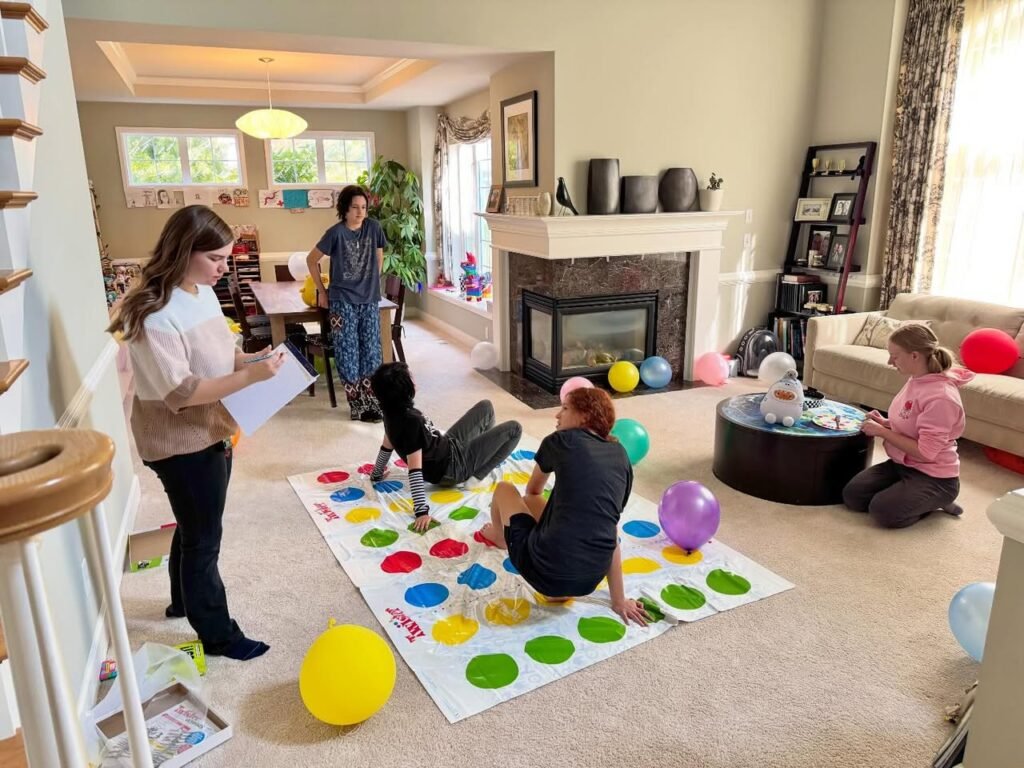 A group of people and a dog playing Twister with giant balls in a spacious living room. The room features a fireplace, large windows, and casual furniture.