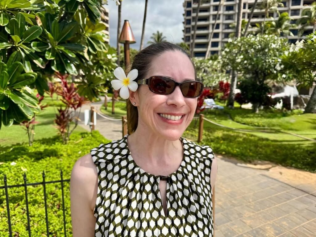 A traveler wearing sunglasses and a floral hairpiece enjoys a sunny day in a tropical setting. Lush green foliage and a spiral tower building characterize the scenic background.