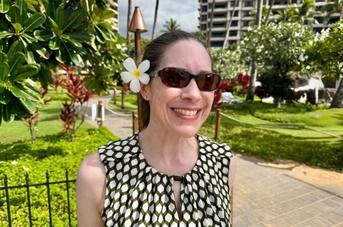 A traveler wearing sunglasses and a floral hairpiece enjoys a sunny day in a tropical setting. Lush green foliage and a spiral tower building characterize the scenic background.