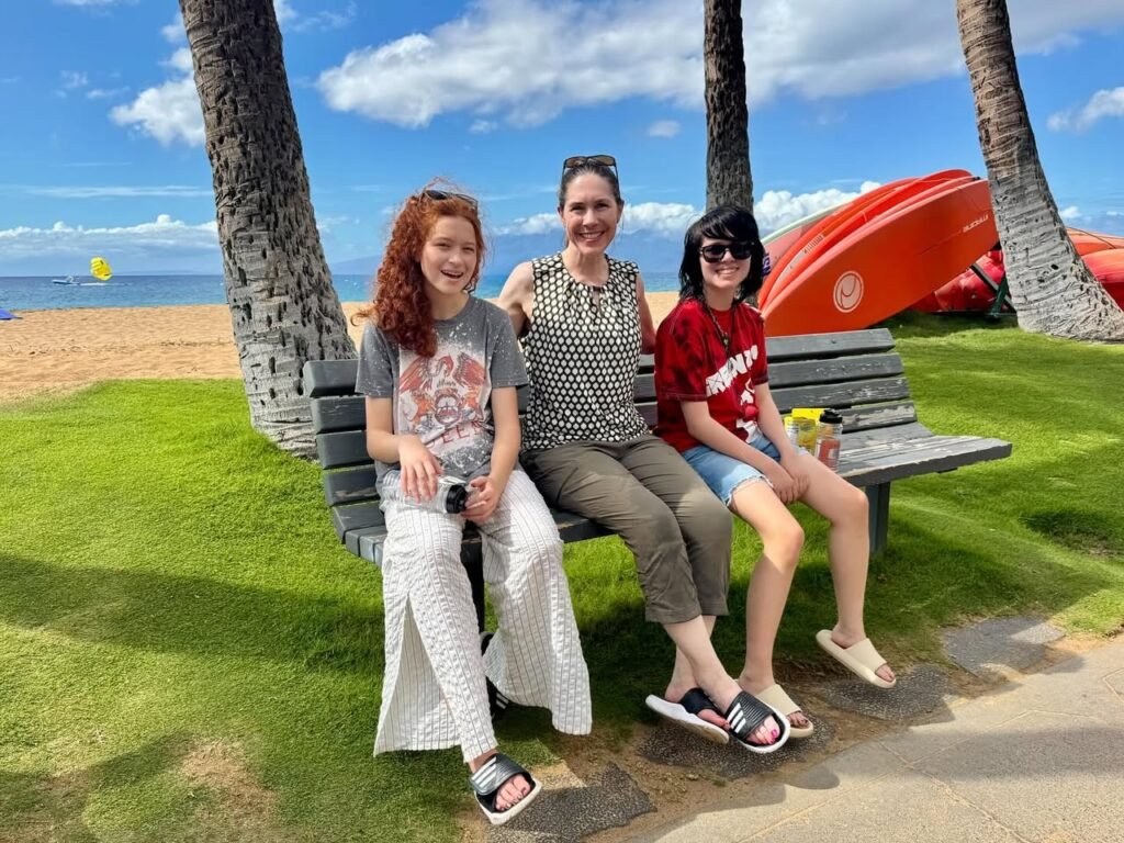 A family seated on a bench at a sunny beach with palm trees in the background. A red lifeguard stand is visible nearby.