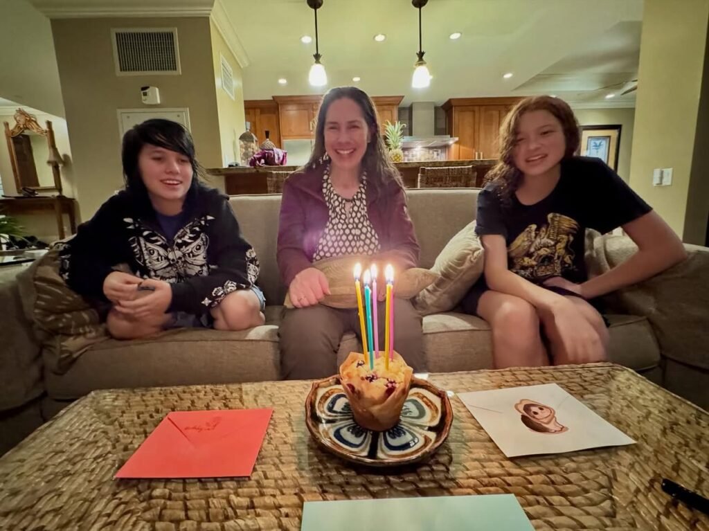 Three people sit on a sofa enjoying a birthday celebration with lit candles in the background.