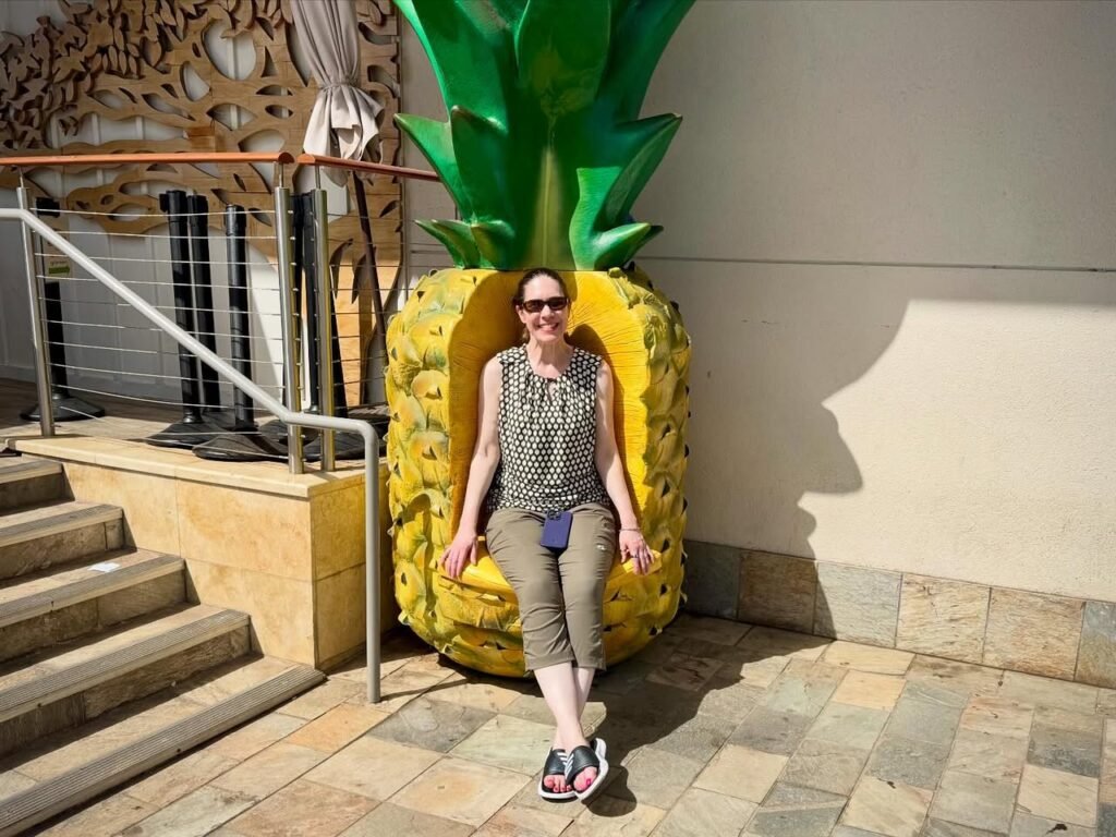 A person takes a fun selfie in a large yellow pineapple sculpture chair outdoors.