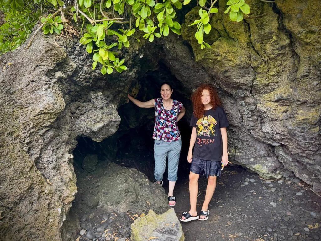 Two people standing in front of a dark rock cave entrance with green foliage overhead. The scene is set within a rugged natural formation.