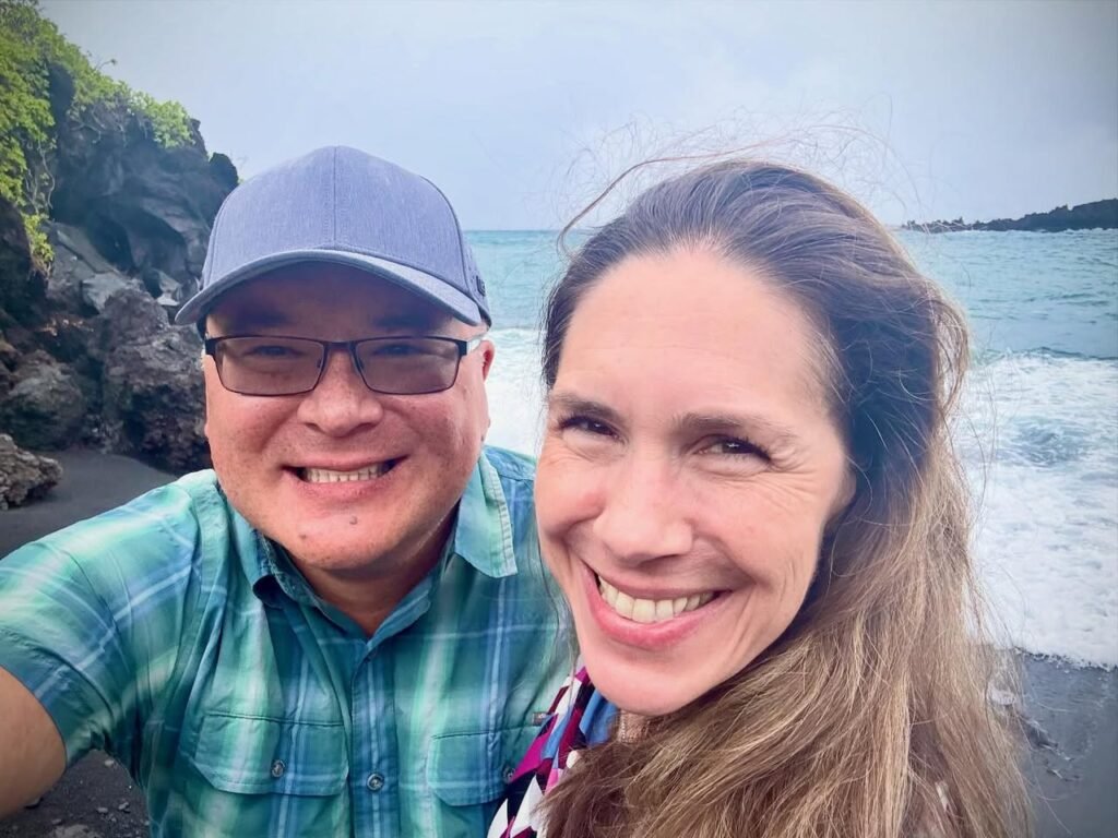 A couple taking a selfie on a black sand beach with a scenic ocean view in Hana, Hawaii.