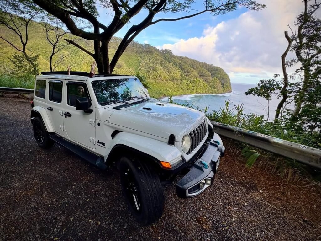 A white Jeep parked on a gravel pullout overlooking a lush coastal landscape.