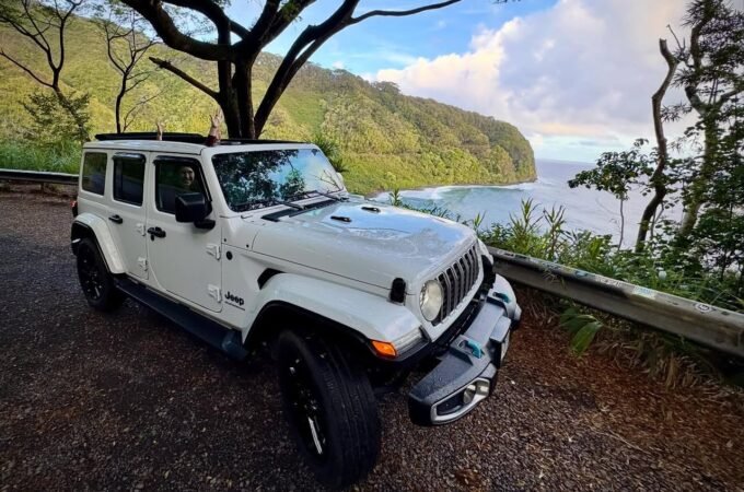 A white Jeep parked on a gravel pullout overlooking a lush coastal landscape.