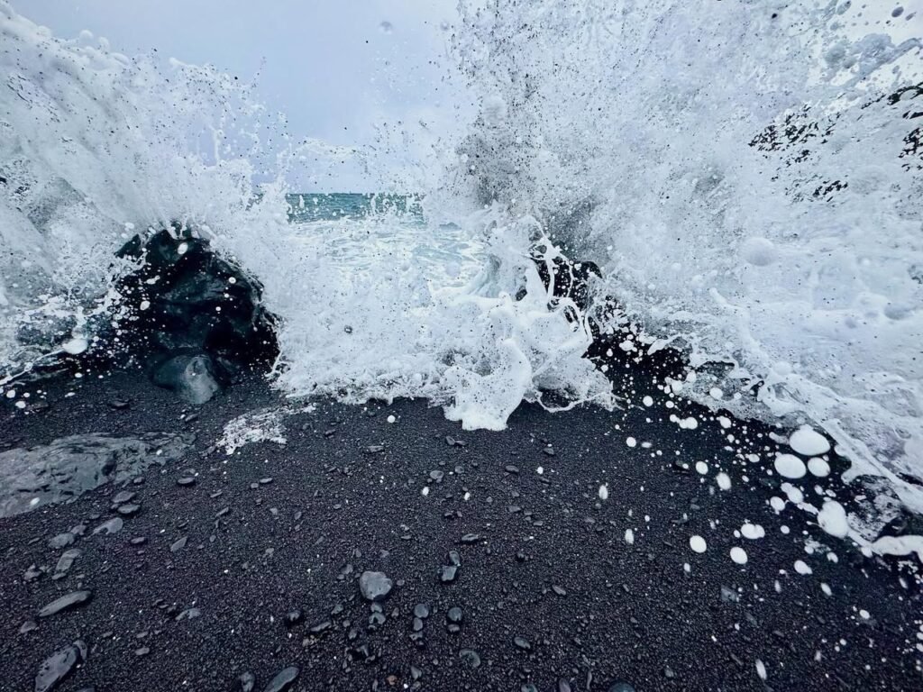 Powerful ocean waves crashing against a black volcanic sand beach. White foam contrasts with dark stones and rocks.