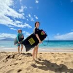 Hosmers Grove Birding Two people holding paddleboards on a sandy beach under a bright blue sky.