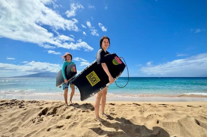 Two people holding paddleboards on a sandy beach under a bright blue sky.